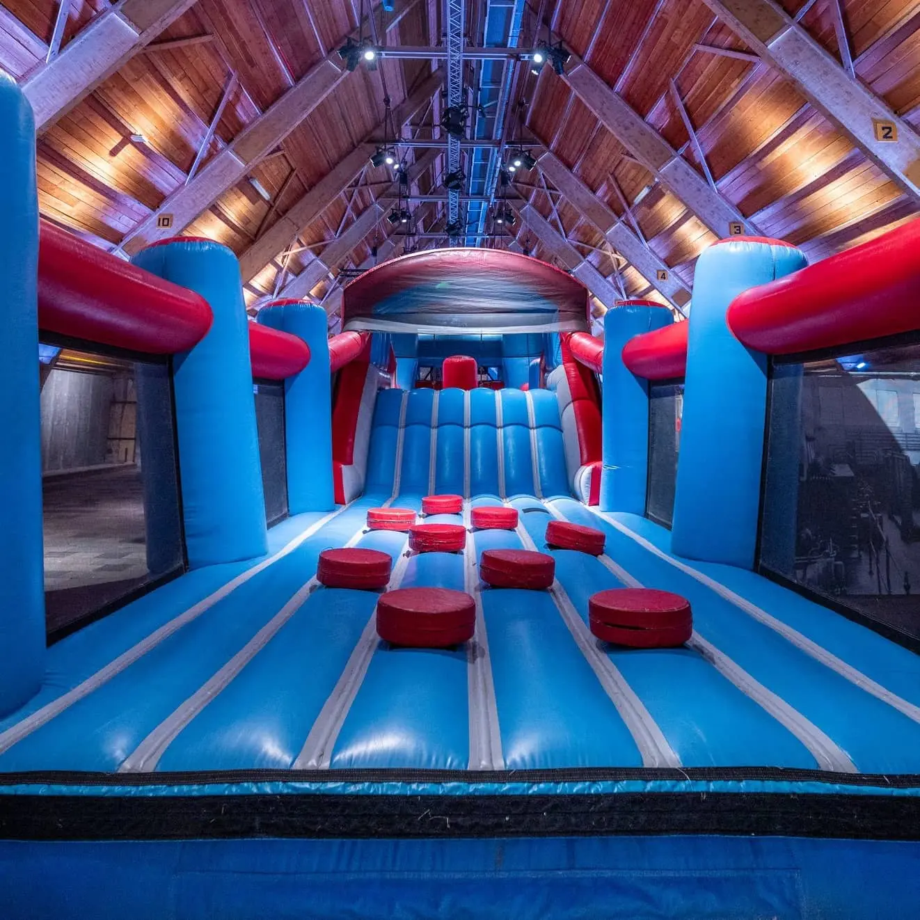 Blauwe en rode indoor stormbaan met zachte hindernissen in een grote houten hal met hoge balken en sfeerverlichting.